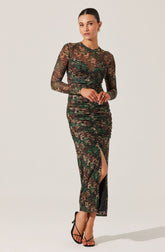 A woman is wearing a long-sleeved, full-length dress made of sheer, floral-printed fabric. The dress features a gathered, ruched design around the waist and hips, creating a fitted silhouette. It has a high slit up one leg, revealing part of her leg and black high-heeled sandals. The floral pattern includes shades of green, brown, and black.